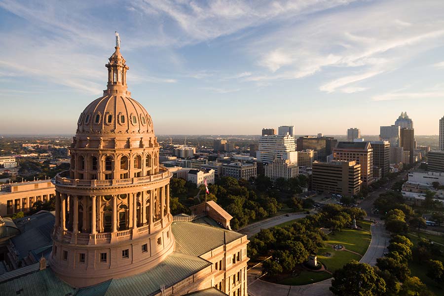 About Us - Ornate Texas State Capitol Building Shining at Sunset, With Green City Parks, Flags Flying, and Tall Buildings Below